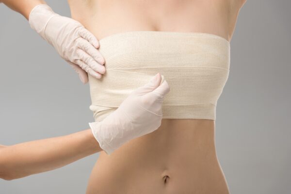 partial view of plastic surgeon in latex gloves and patient in breast bandage isolated on grey