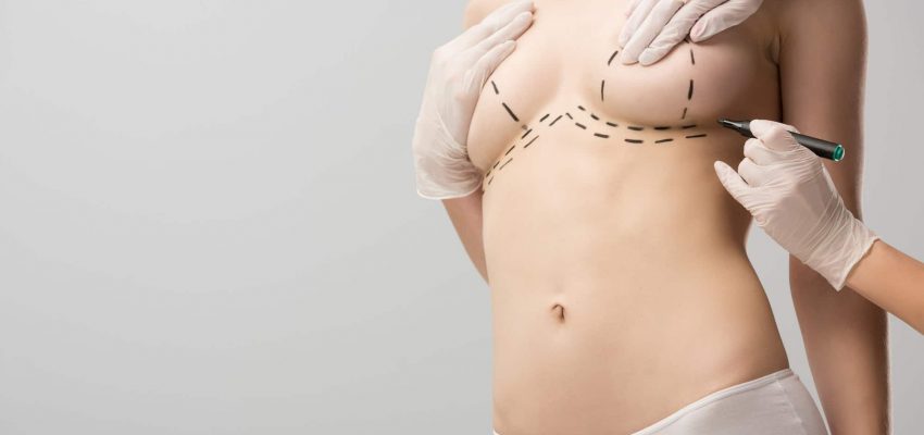 cropped view of plastic surgeon in latex gloves making marks on breast isolated on grey